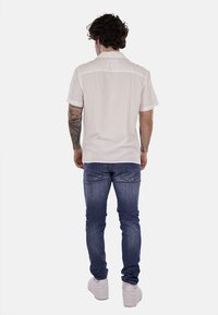Cammicia bianca a maniche corte, realizzata in tessuto leggero, con un taglio dritto e un design semplice sul retro, abbinata a jeans blu scuro e scarpe bianche.