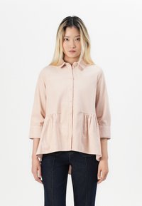 Blusa rosa chiaro, con bottoni e colletto, maniche a tre quarti e vita arricciata. Tessuto testurizzato e orlo arrotondato. Abbinata a jeans scuri.