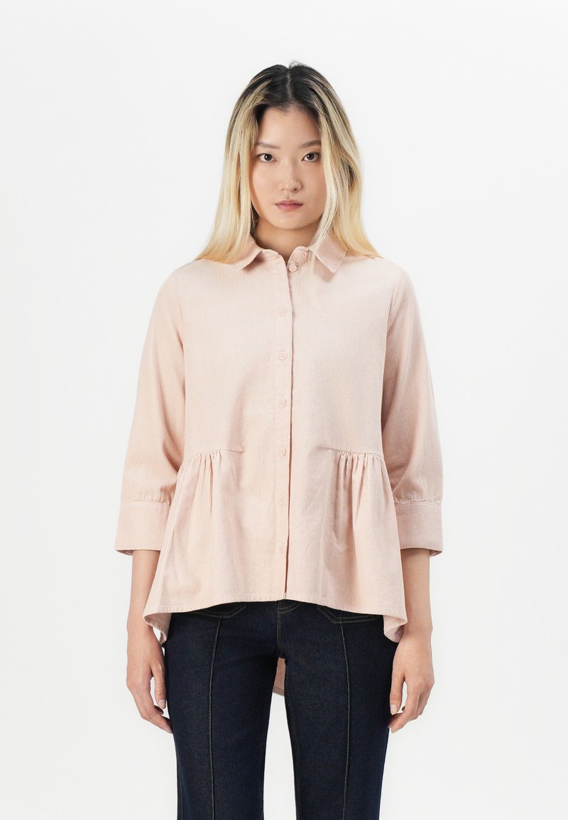 Blusa rosa chiaro, con bottoni e colletto, maniche a tre quarti e vita arricciata. Tessuto testurizzato e orlo arrotondato. Abbinata a jeans scuri.