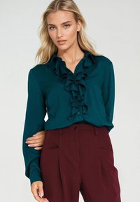 Turkusowa bluzka z falbaną z przodu, zapinana na guziki, z długimi rękawami, zestawiona z bordowymi spodniami z bocznymi kieszeniami i gładką fakturą.