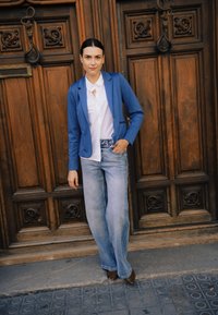 Blazer bleu porté sur une chemise blanche avec un détail en forme de trou de serrure, associé à un jean large bleu clair et des chaussures marron, sur fond de porte en bois.
