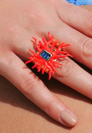 Bague en forme de corail rouge avec une pierre rectangulaire bleue au centre. Le design est complexe avec des accents texturés sur un anneau en or.