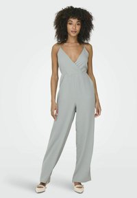 Mono gris claro con escote en forma de V y tirantes finos. Piernas anchas y holgadas con una textura suave. Combinado con bailarinas de color claro.