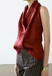 Haut sans manches en lin rouge profond à col drapé, associé à un pantalon taille haute marron à fines rayures et un bracelet manchette doré texturé.