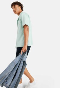 Cămașă verde mentă cu mânecă scurtă și un buzunar pe piept, purtată cu pantaloni scurți negri și adidași albi. Ținând o geacă din denim albastru.
