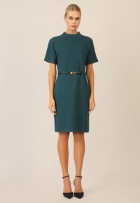 Vestido de punto en tono teal con mangas cortas, cuello alto y un cinturón negro con hebilla dorada. Combinado con tacones negros de punta afilada.