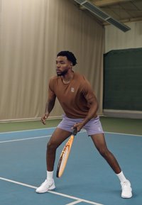 Tennis spiller i en brun kortermet skjorte og lyse lilla shorts, som holder en racket med et oransje og svart design, på en blå bane.