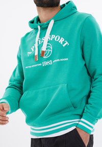 Felpa turchese con coulisse bianche e stampa rétro. Presenta una tasca a marsupio e polsini a righe. Tessuto morbido, vestibilità casual.
