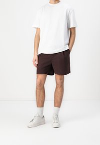 Valkoinen lyhythihainen t-paita, tummat ruskeat shortsit, harmaat tekstuuriset sukat ja valkoiset lenkkitossut. Rentoa asustusta yksinkertaisilla, siisteillä linjoilla.