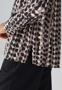 Kurzarmpf blouse aus braunem Stoff mit großen Schwarz-Weiß-Punktemustern, geradem Saum und seitlichen Schlitzen für mehr Komfort.