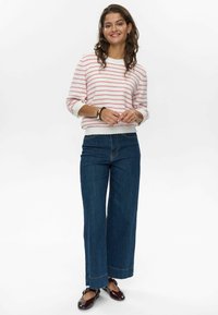 Fehér és rózsaszín csíkos kötött pulóver, fodros szegéllyel, széles lábú sötétkék farmernadrággal és barna cipővel párosítva.