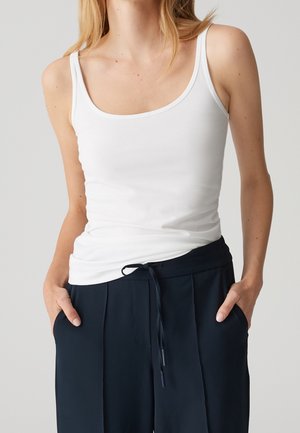 Personne portant un débardeur blanc ajusté sans manches et un pantalon noir ample à cordon, les mains dans les poches, sur un fond uni.