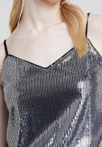 Femme portant un top en camisole noir et argent à paillettes avec de fines bretelles et des cheveux blonds clairs sur une épaule.