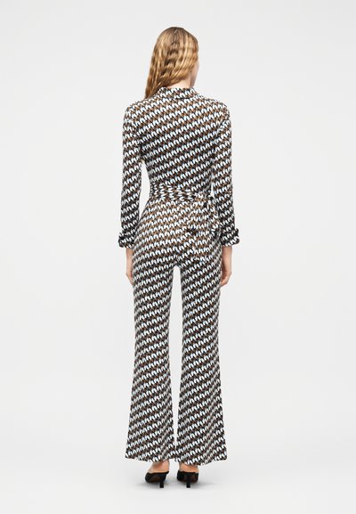 Γυναίκα με μακριά κυματιστά ξανθά μαλλιά φορώντας εφαρμοστή ολόσωμη φόρμα με καφέ, μαύρο και λευκό μοτίβο, με καμπάνα στα πόδια και μαύρα παπούτσια με τακούνια, γυρισμένη προς τα πίσω.