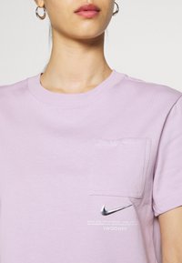 Lila bomulls-t-shirt med rund halsringning, korta ärmar, en vänster bröstficka och en metallic Nike swoosh-logga tryckt under fickan.