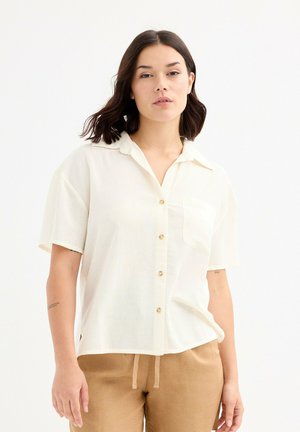 Camisa crema de botón, con cuello, mangas cortas y un bolsillo frontal. Presenta botones dorados y una tela ligera y texturizada.