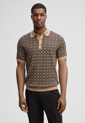 Uomo che indossa una polo a maniche corte beige e nera con motivo a fantasia e colletto con bottoni, in piedi davanti a uno sfondo chiaro uniforme.