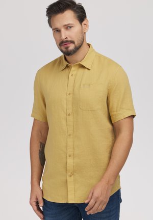 Mężczyzna z brodą, ubrany w żółtą koszulę z krótkim rękawem w kolorze musztardowym i niebieskie dżinsy, stojący na jednolitym jasnym tle.