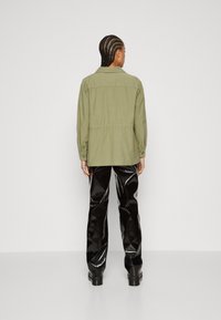 Khaki jacket with a cinched waist and buttoned cuffs, paired with glossy black flare pants and sturdy black boots, shot from behind.