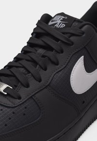 Prim-plan cu pantof sport Nike Air negru, cu panouri texturate, șireturi groase negre și sigla albă Nike pe lateral.