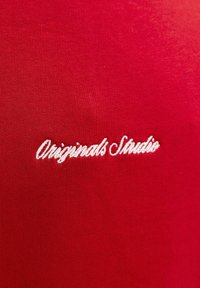 Texte blanc en cursive "Originals Studio" brodé sur fond en tissu rouge.