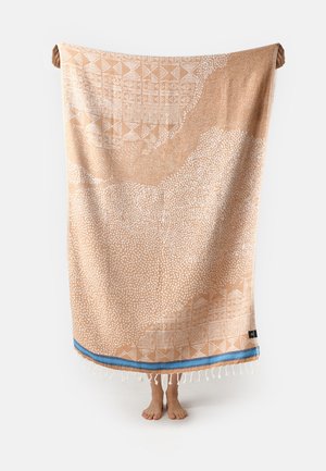 Persona che tiene un grande tessuto beige e bianco con motivo, banda blu e frange, piedi visibili sotto su sfondo bianco.