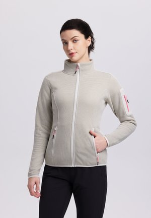 Woman wearing a light gray textured zip-up jacket with high collar, hand in pocket, and black pants, standing against plain background.