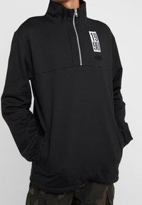Czarny sweatshirt z ćwierć zamkiem, wykonany z miękkiego materiału, z długimi rękawami, zamkiem w kołnierzu oraz detalem logo na przodzie z numerami "1990."