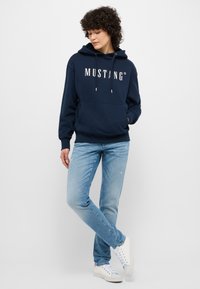 Felpa navy con logo bianco "MUSTANG", cappuccio con cordino e tasca frontale. Abbinata a jeans chiari strappati e sneakers bianche.