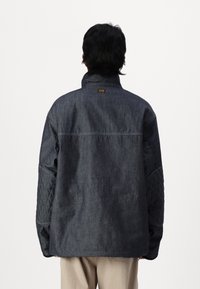Temno denim jakna s kapicami in visokim ovratnikom. Ima vidno šivanje in majhno oznako blagovne znamke na vratu. Ohlapen kroj.