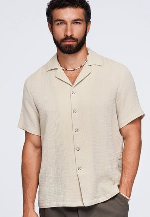 Homme avec une barbe portant une chemise beige à manches courtes boutonnée et un collier de perles, debout avec une main dans sa poche sur un fond uni.