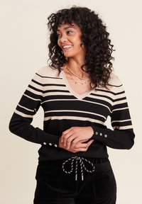 Schwarzer V-Ausschnitt-Pullover mit metallisch goldenen horizontalen Streifen, mit Knopf-Details an den Ärmeln und einer gerippten Textur. Kombiniert mit dunklen Hosen.