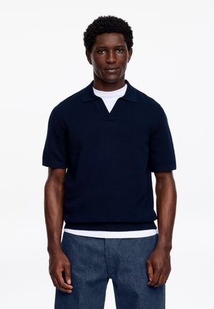 Uomo che indossa un maglione a maniche corte blu navy sopra una camicia bianca e pantaloni blu, in piedi contro uno sfondo bianco semplice.