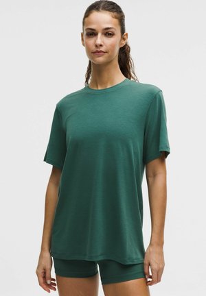 Femeie cu păr închis la culoare prins în coadă de cal, purtând un tricou larg cu mâneci scurte de culoare verde închis și pantaloni scurți mulați de aceeași culoare, stând pe un fundal deschis.