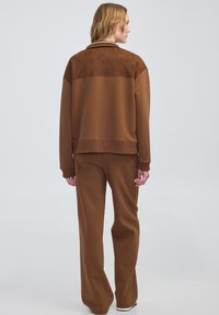 Tuta marrone con una parte superiore in suede texturizzato e una parte inferiore in tessuto liscio; presenta polsini a costine e una vestibilità rilassata.