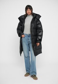 Chaqueta de plumas larga y negra con un alto cuello y acabado brillante. Combinada con una camiseta gris, jeans azul claro y zapatillas verdes.