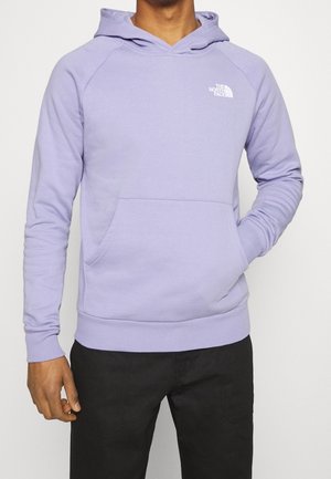 Mężczyzna w jasnofioletowej bluzie The North Face z przednią kieszenią i czarnych spodniach, ręka spoczywa w kieszeni, na jednolitym tle.