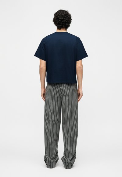 Osoba s kudrnatými tmavými vlasy stojí zády, má na sobě tmavě modré triko s krátkým rukávem a volné šedé kalhoty s vertikálními černými pruhy.