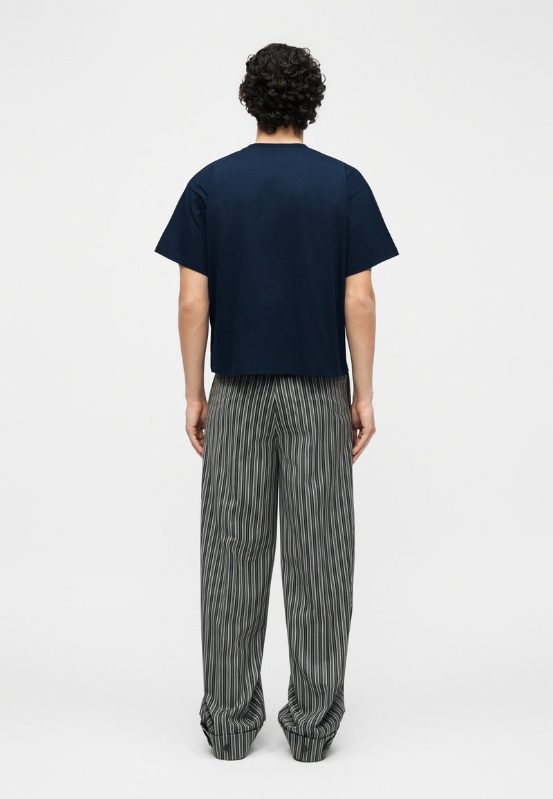 Persona con capelli ricci e scuri vista di spalle, indossa una maglietta blu scuro a maniche corte e pantaloni larghi grigi con strisce verticali nere.