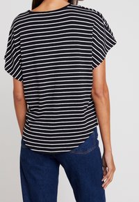 Top de manga corta a rayas blancas y negras con escote redondeado y aberturaslaterales, combinado con pantalones de mezclilla azules. Se puede ver la textura suave del tejido.