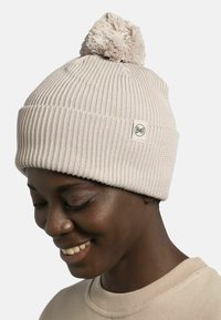 Gorro de punto beige claro con un pom-pom en la parte superior, que presenta una textura acanalada y una pequeña etiqueta circular del logo de Buff en el lado.