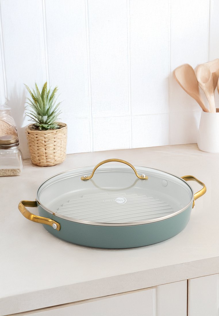 Poêle ronde avec un extérieur vert léger, un couvercle en verre et des poignées dorées ; présente un intérieur strié et un bord argenté.