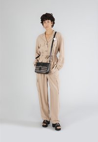 Beige, långärmad jumpsuit med knyt i midjan, knappstängning och fickor; matchad med en svart axelväska med flätad detalj och metalliska accenter.