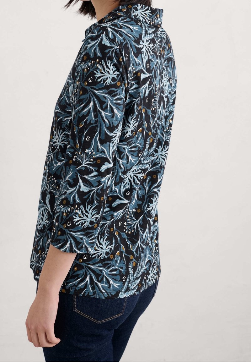 Femme portant un chemisier noir à motifs de feuilles bleues et blanches avec des manches trois-quarts, assorti à un jean bleu foncé.