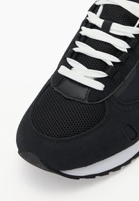 Chaussure de sport noire en matériaux mesh et suédé, avec des lacets blancs, une surface texturée et une semelle en caoutchouc noire avec une semelle intermédiaire blanche.