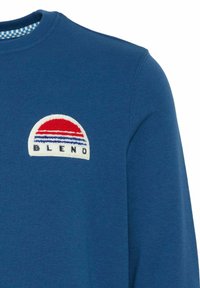 Felpa blu con un logo ricamato tondo che presenta strisce rosse e blu e la parola "BLEND" su uno sfondo bianco.