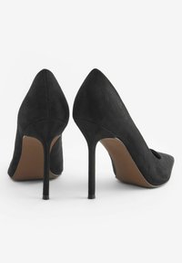 Scarpe con tacco alto in suede nero con punta affilata e dettaglio a taglio, dotate di un sottile tacco a stiletto e una suola liscia marrone.