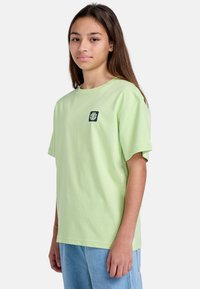 Camiseta de algodón verde claro con cuello redondo y mangas cortas; presenta un pequeño logo azul oscuro en la parte izquierda del pecho.