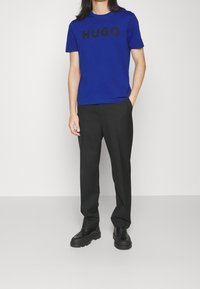 Modré bavlněné tričko s nápisem "HUGO" v černém písmu, doplněné o černé kalhoty a černé platformové boty, vše na neutrálním pozadí.