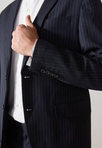 Mornarsko prugasta sako sa četiri dugmeta na manžetama, jednim džepom na prsima i kontrastnom bijelom košuljom ispod.